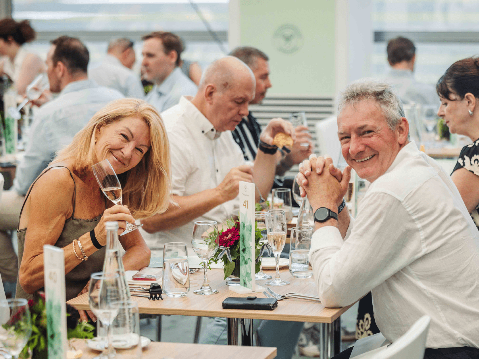A couple smiling at their table in The Lawn holding a glass of Champagne at Wimbledon 2025